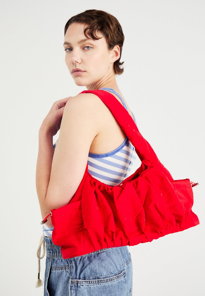 Bolsa de tela roja con textura fruncida, correa ajustable y herrajes plateados. El modelo lleva una camiseta de tirantes a rayas y pantalones cortos de mezclilla.