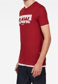 Red cotton t-shirt with a crew neckline, featuring a prominent white "RAW." logo across the chest. Short sleeves, slim fit design.