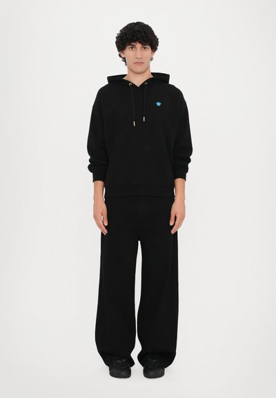 Felpa nera con cappuccio e coulisse dorata, abbinata a pantaloni neri larghi e scarpe nere, su uno sfondo bianco.