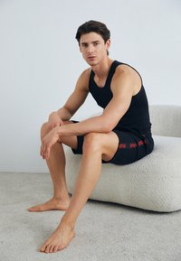 Črni top brez rokavov in črnske kratke hlače z rdečim besedilom, sedeč na teksturirani, svetli oblazinjeni površini, bos na mehkem tleh.