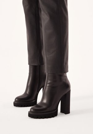 Schwarze kniehohe Stiefel mit hohem Absatz und klobigen Plateau-Sohlen, getragen mit schwarzen Lederhosen.