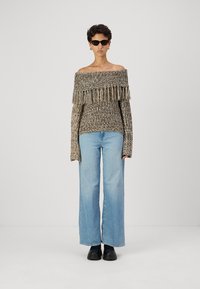 Pulover tricotat off-the-shoulder de culoare maronie, cu detalii din franjuri, asortat cu blugi evazați de culoare bleu deschis și pantofi negri.