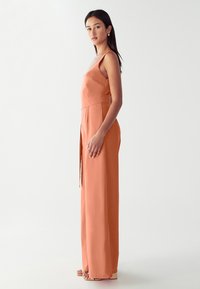 Žena stojící v profilu, oblečená v bezrukávovém korálovém overal a béžových platformových sandálech, na jednobarevném světlém pozadí.