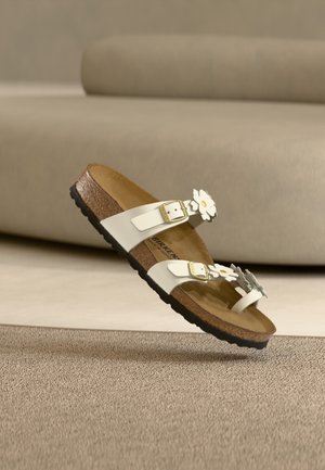Άσπρο σανδάλι Birkenstock με φελλό στη σόλα, δύο λουράκια με αγκράφες και διακοσμητικά λουλούδια μαργαρίτας πάνω σε μπεζ χαλί.