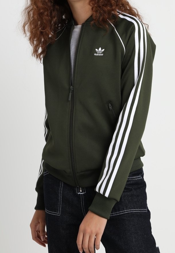 Mörkgrön zip-up jacka i slätt tyg, med vita tre-stripes detaljer på ärmarna och en liten logotyp på bröstet.