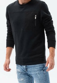 Sudadera negra de manga larga, con un panel frontal texturizado y un bolsillo en el pecho con cremallera. Combinada con jeans desgastados de color azul claro.
