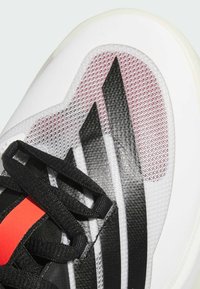 Scarpa sportiva bianca con accenti neri e rossi, texture in rete, strisce prominenti e lacci neri piatti. Design con punta arrotondata.