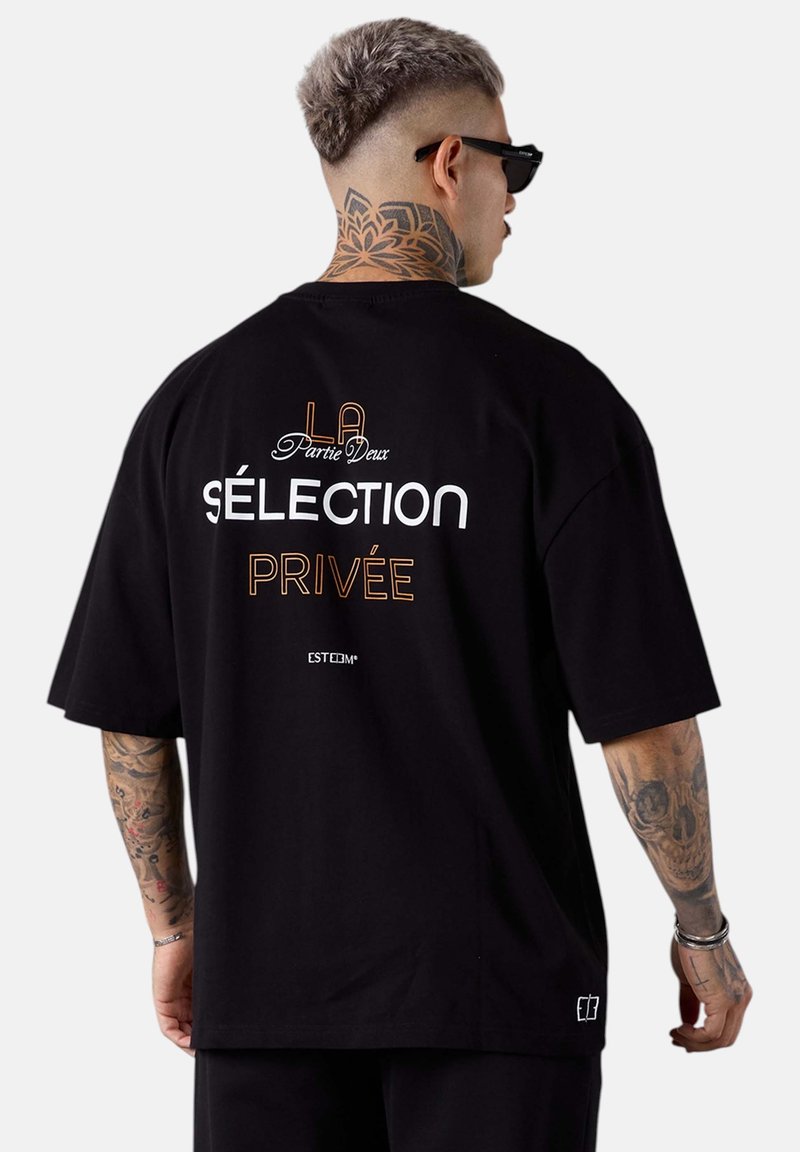 Schwarzes, übergroßes T-Shirt mit weißem und orangefarbenem Text auf dem Rücken; bemerkenswerte Elemente sind ein blumiges Tattoo am Hals und kurze Ärmel.