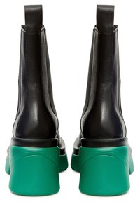 Botas negras de cuero hasta el tobillo con suelas de goma brillante verde y tiradores, mostradas desde la parte de atrás sobre un fondo blanco.