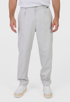 Mężczyzna ubrany w jasnoszare plisowane spodnie i białe sneakersy, stojący z luźno opuszczonymi ramionami na jednolitym tle.