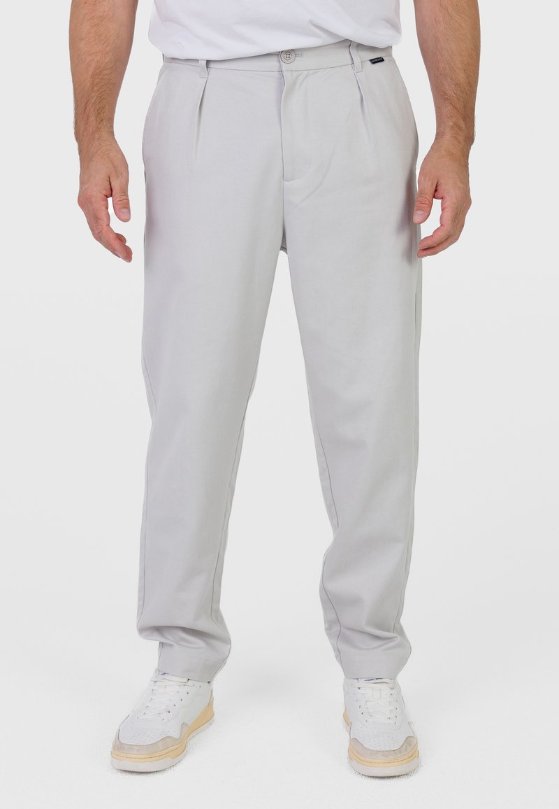 Homme portant un pantalon plissé gris clair et des baskets blanches, debout avec les bras détendus le long du corps, sur un fond uni.
