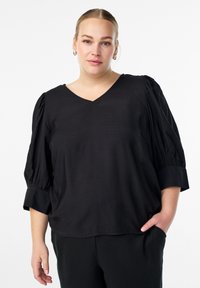 Vrouw met blond haar in een lage bun, draagt zilveren creolen, een zwarte V-hals blouse met puffmouwen en zwarte broek, met één hand in de zak.