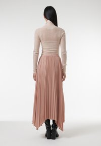 Lichtroze geplooide rok met een asymmetrische zoom, gecombineerd met een getailleerd, doorzichtig long-sleeve top. Model draagt zwarte kniehoge laarzen.