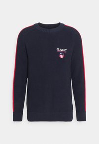 Marineblauer Strickpullover mit roten Seitenstreifen, geripptem Kragen und Bündchen. Mit gesticktem "GANT"-Logo und Wappen auf der Brust.