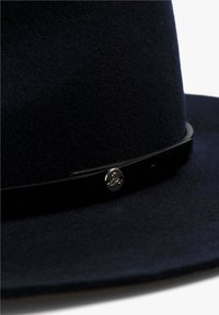 Chapeau en feutre marine avec un large bord, doté d'un bandana en cuir noir et d'un accent en forme de crâne argenté à l'avant. Texture douce et forme structurée.