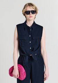 Persona che indossa una camicia smanicata button-up di un blu navy scuro e pantaloni, occhiali da sole neri, tenendo una borsa clutch intrecciata di un brillante colore rosa nella mano sinistra.