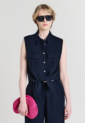 Person, die ein dunkeln navy-blaues ärmelloses Hemd mit Knöpfen und eine Hose trägt, schwarze Sonnenbrille aufhat und in der linken Hand eine leuchtend pinkfarbene geflochtene Clutch hält.