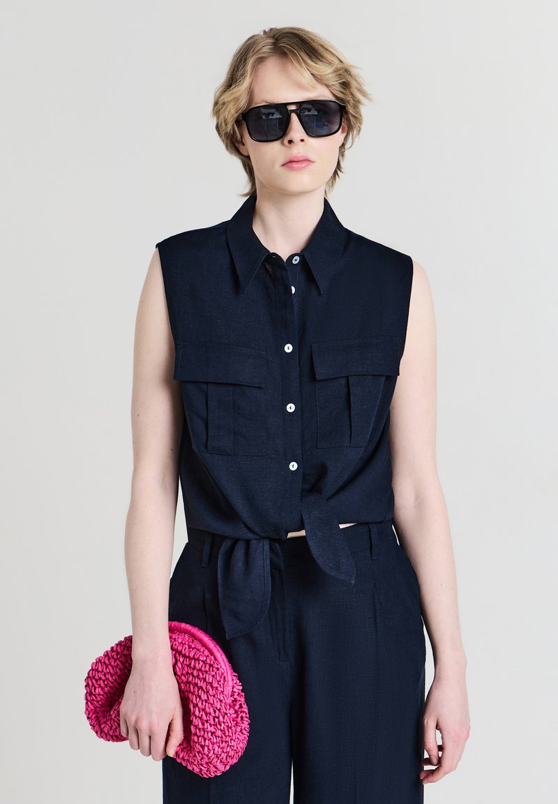 Persona che indossa una camicia smanicata button-up di un blu navy scuro e pantaloni, occhiali da sole neri, tenendo una borsa clutch intrecciata di un brillante colore rosa nella mano sinistra.