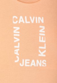 Camiseta color durazno con una textura suave que presenta letras blancas en bloque que dicen "CALVIN KLEIN JEANS" dispuestas en un diseño rectangular.