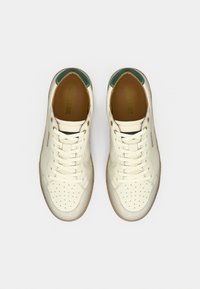 Ζευγάρι από λευκά leather sneakers με διάτρητα στα δάχτυλα, κρεμ κορδόνια, πράσινες λεπτομέρειες στην φτέρνα και καφέ εσωτερικά πέλματα σε λευκό φόντο.