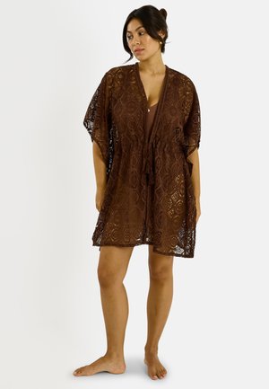 Mujer con el cabello oscuro recogido en un moño, vestida con un vestido marrón hasta la rodilla con patrón de encaje, de pie descalza, mirando hacia un lado.