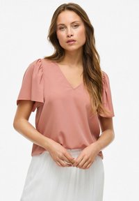 Blusa rosa a V con maniche corte a bufalo. Tessuto morbido e leggero, design leggermente drappeggiato. Abbinata a una gonna bianca plissettata.