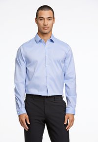Camicia azzurro chiaro con bottoni, realizzata in tessuto morbido, dotata di colletto classico, maniche lunghe e bottoni sui polsini. Abbinata a pantaloni scuri.