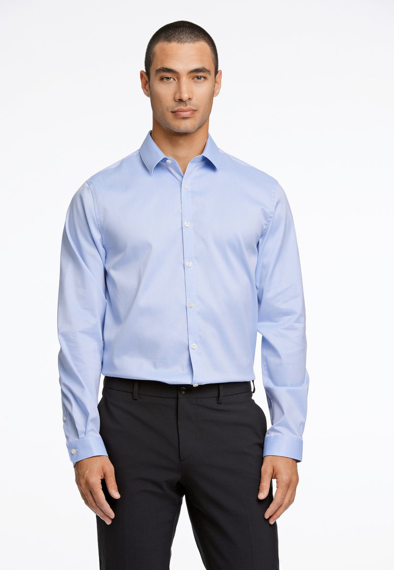 Camicia azzurro chiaro con bottoni, realizzata in tessuto morbido, dotata di colletto classico, maniche lunghe e bottoni sui polsini. Abbinata a pantaloni scuri.