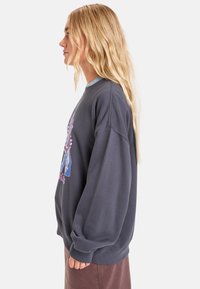 Sudadera gris oscuro con cuello redondo, corte oversize y mangas largas. Presenta un estampado gráfico colorido en la parte frontal.