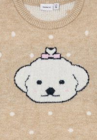Beige strikkegenser med hvite prikker og en brodert hundefjes med en rosa sløyfe, laget av mykt, strukturert garn.