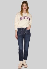 Jeune femme souriante, portant un sweat-shirt crème avec l'inscription "BOSTON 1980", un jean bleu foncé, des chaussettes crème et des baskets beige avec des détails bordeaux et noirs.