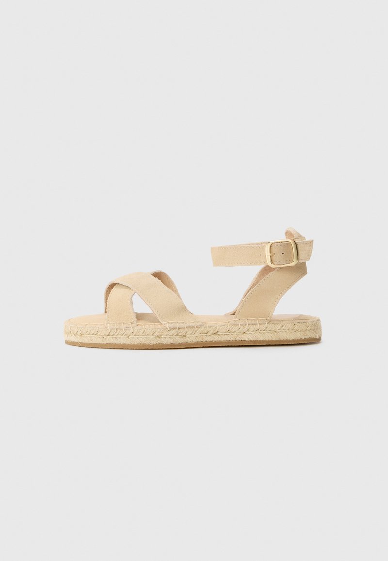 Tan espadrille sandals featuring a woven jute sole, two crisscross straps, and an adjustable ankle strap with a gold buckle accent.