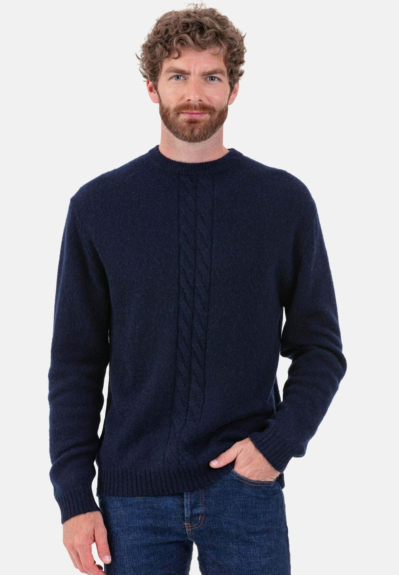 Pull en maille bleu marine fabriqué en matériau doux, avec un motif torsadé central et des poignets et ourlet côtelés. Le modèle se tient les mains dans les poches.