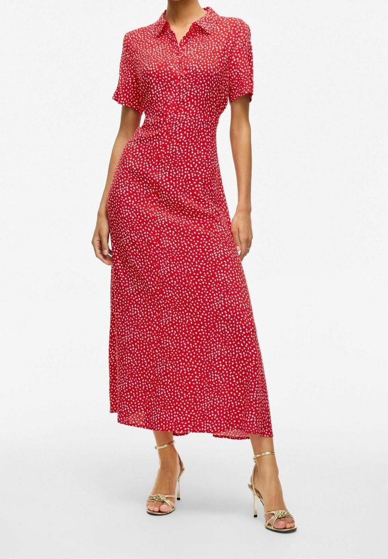 Robe maxi rouge à manches courtes avec un devant boutonné, présentant un motif à pois et une silhouette fluide. Associée à des sandales à talons hauts dorées.