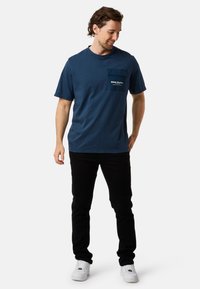 T-paita tummansinisestä puuvillasta, jossa on pyöreä kaula-aukko, lyhyet hihat ja taskupolttomaalaus, jossa on valkoista tekstiä. Yhdistetty mustiin housuihin ja valkoisiin lenkkareihin.