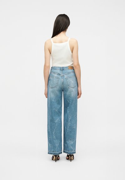 Mujer con cabello largo y oscuro, vistiendo una camiseta blanca sin mangas, vaqueros azul claro de talle alto y pierna ancha con patrones descoloridos, y tacones negros, de pie.