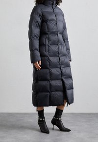Dlouhý černý péřový kabát s vysokým límcem, obdélníkovým prošíváním, předním zipem, bočními kapsami a délkou sahající po kolena.