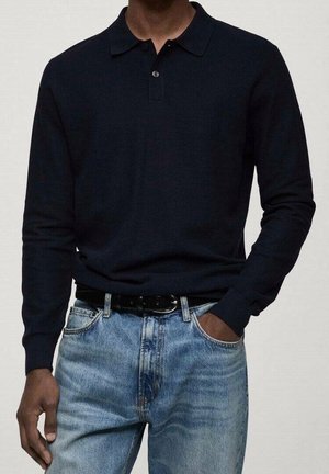 Mann trägt ein schwarzes langärmeliges Polo, in hellblaue Jeans mit schwarzem Gürtel gesteckt, eine Hand in der Tasche.