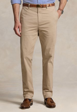 Mies pukeutuu beigenvärisiin housuihin, ruskeaan vyöhön, siniraitaisen paitaan, ruskeisiin tupsulööfereihin ja rannekelloon, seisoo rentoutunein käsin.