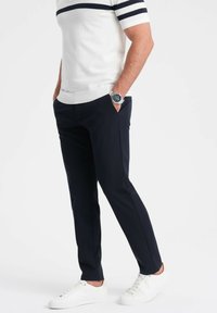 Marineblauwebroek met een slim fit snit, gecombineerd met een witte top met korte mouwen en marineblauwe strepen, en witte sneakers. Horloge om de pols.