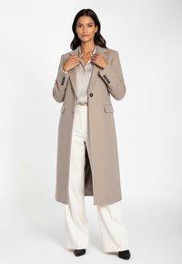 Beige lång kappa med texturerad finish, slag med hack och svarta knappar, kombinerad med en ljus blus och vita vida byxor.