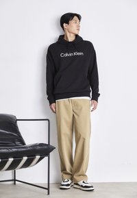 Black hooded sweatshirt with white "Calvin Klein" logo, paired with beige wide-leg trousers and black-and-white sneakers. Minimalist style.