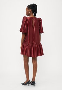 Σατέν μίνι φόρεμα σε μπορντό απόχρωση με φουσκωτά μανίκια, ριγμένο στρίφωμα και λεπτομέρεια κλειδαρότρυπας στην πλάτη. Συνδυάζεται με μαύρες ψηλοτάκουνες γόβες με μυτερή μύτη.