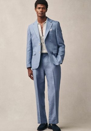 Traje de lino azul claro con un corte estructurado, chaqueta de un solo botón y pantalones a juego; combinado con una camisa clara y zapatos oscuros.