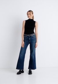 Μαύρο αμάνικο τοπ με διακοσμητικά κουμπιά στο πλάι, σε συνδυασμό με καμπάνες σκούρες μπλε τζιν και χοντρές μαύρες μπότες. Απλό λευκό φόντο.