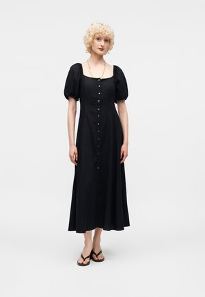 Γυναίκα με σγουρά ξανθά μαλλιά φοράει μαύρο μίντι φόρεμα με κουμπιά και μανίκια φουσκωτά και μαύρα σανδάλια, στέκεται μπροστά σε ένα λευκό φόντο.