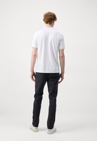 Witte poloshirt met korte mouwen, een gladde textuur, gedetailleerde kraag; gecombineerd met donkere broek en witte sneakers. Neutrale achtergrond.