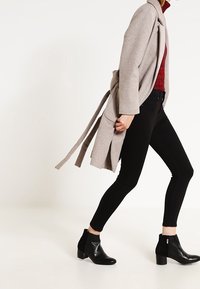 Beige wikkeljas gemaakt van zachte stof met een losse pasvorm, gedragen over een rode top en zwarte skinny jeans, gecombineerd met zwarte enkellaarsjes.