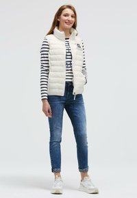 Weißer, plüschiger Weste mit hohem Kragen, die über einem gestreiften Langarmshirt getragen wird. Dunkelblaue Jeans mit hochgekrempelten Säumen. Weiße Sportschuhe.