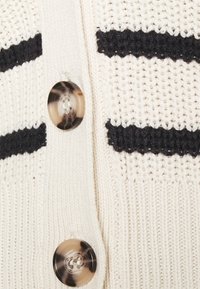 Krämmig stickad cardigan med svarta ränder, stora sköldpaddsknappar, texturerad tyg och ribbad fåll.